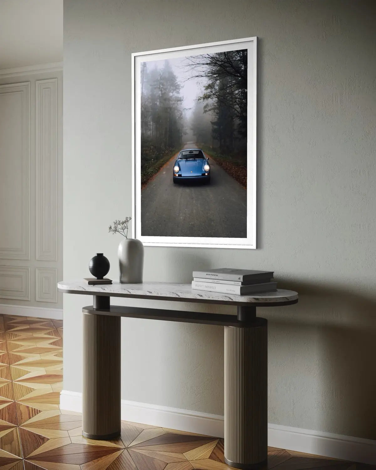 A framed vertical art print depicting a classic blue car driving through a misty forest road, displayed on a light gray wall above a modern console table in a styled interior.