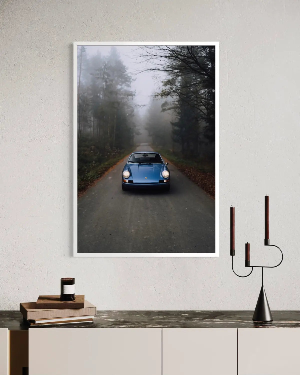 A vertical framed wall art print showing a blue classic car driving down a foggy forest road, displayed on a light wall above a console table with books and a candle holder.