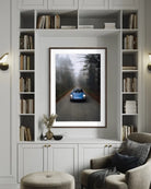 A framed art print depicting a blue classic car driving down a misty forest road, displayed on a white built-in bookshelf above a gray armchair in a styled living room.