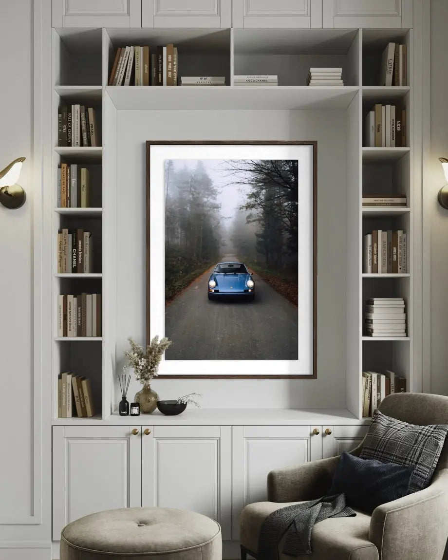 A framed art print depicting a blue classic car driving down a misty forest road, displayed on a white built-in bookshelf above a gray armchair in a styled living room.