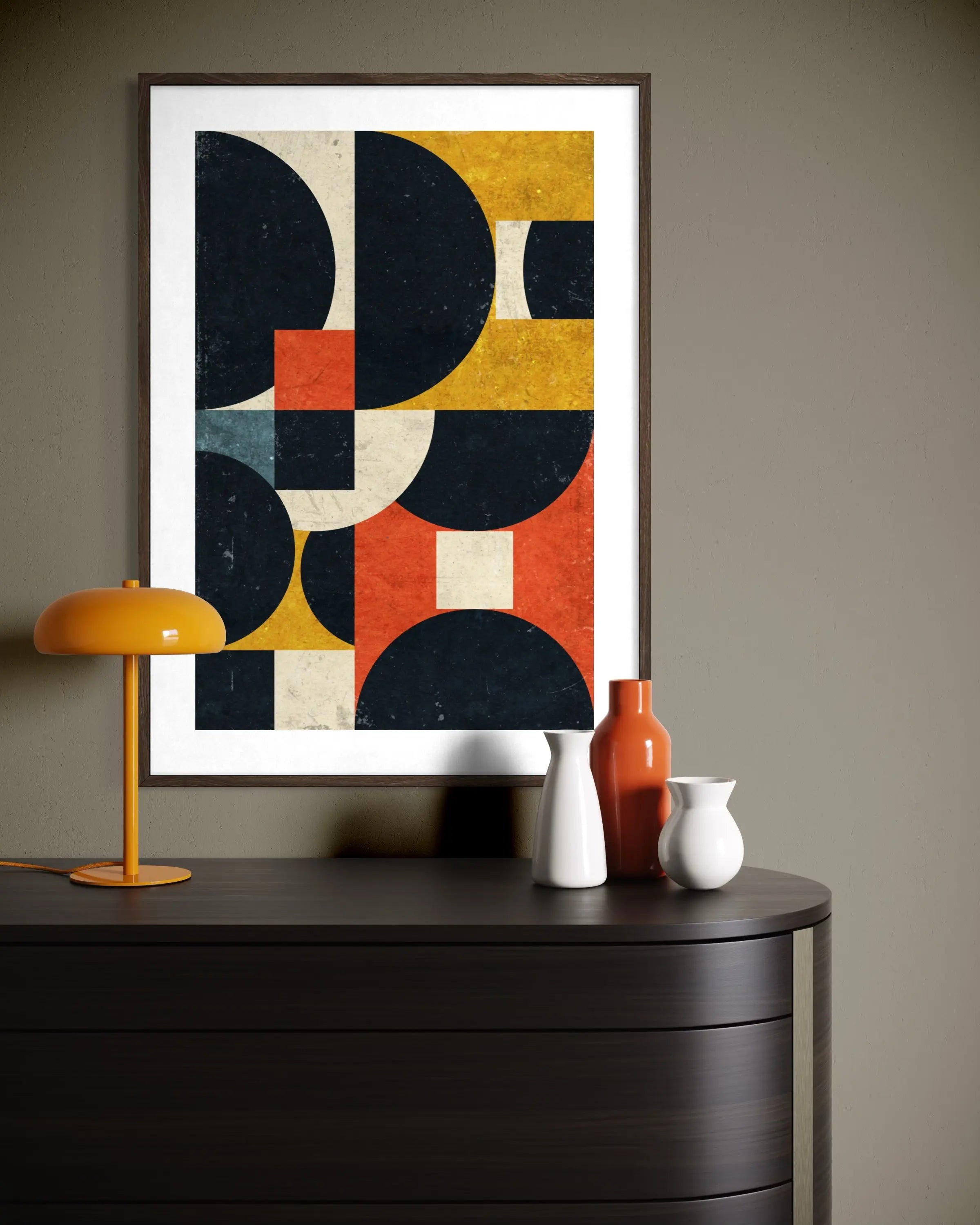 A framed abstract geometric wall art print with bold black, mustard yellow, terracotta orange, and cream shapes, displayed above a dark wood console table with decorative vases and a yellow lamp.
