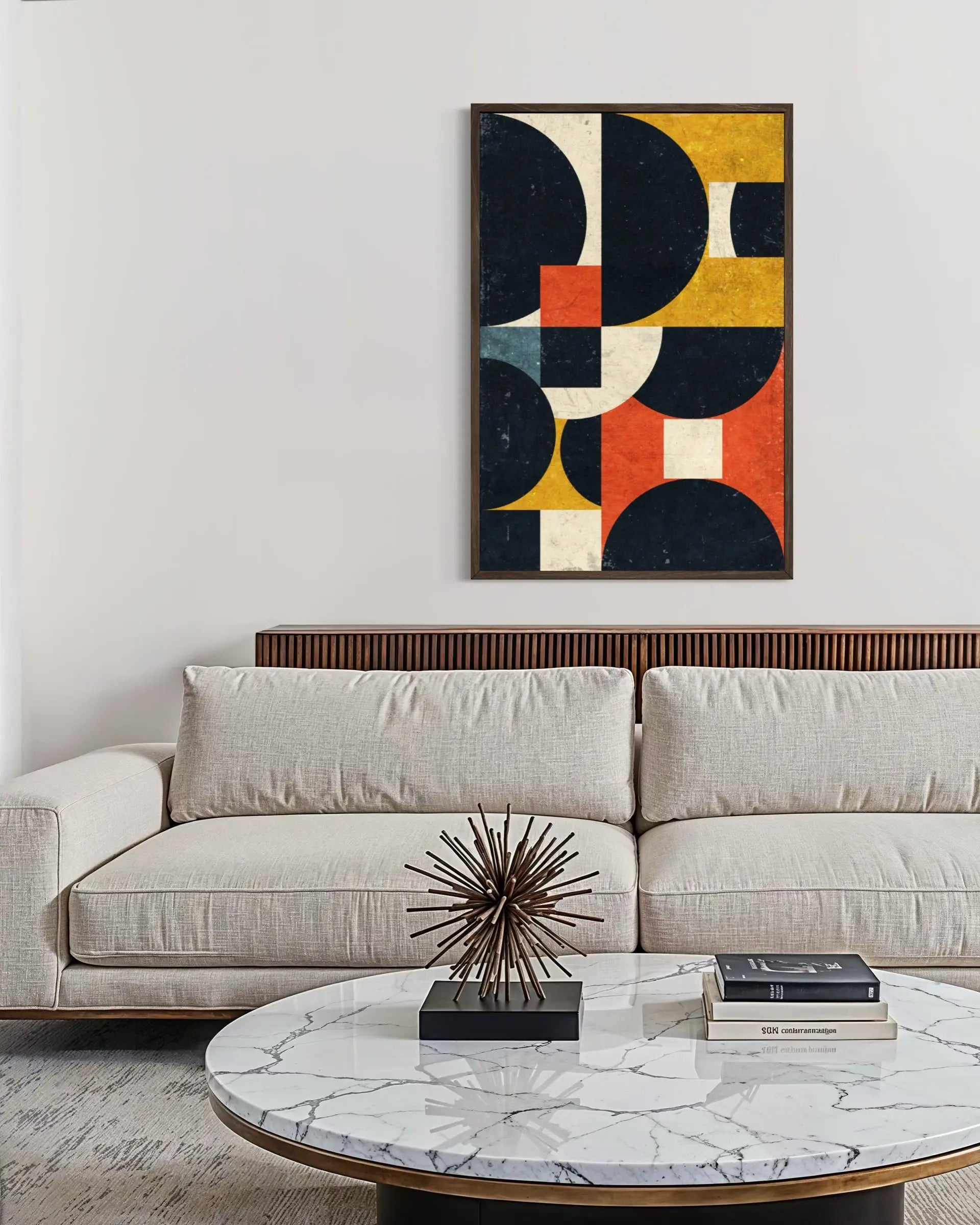 A framed abstract geometric wall art print with navy, mustard yellow, orange, cream, and gray shapes, hanging above a light beige sofa in a modern living room setting.