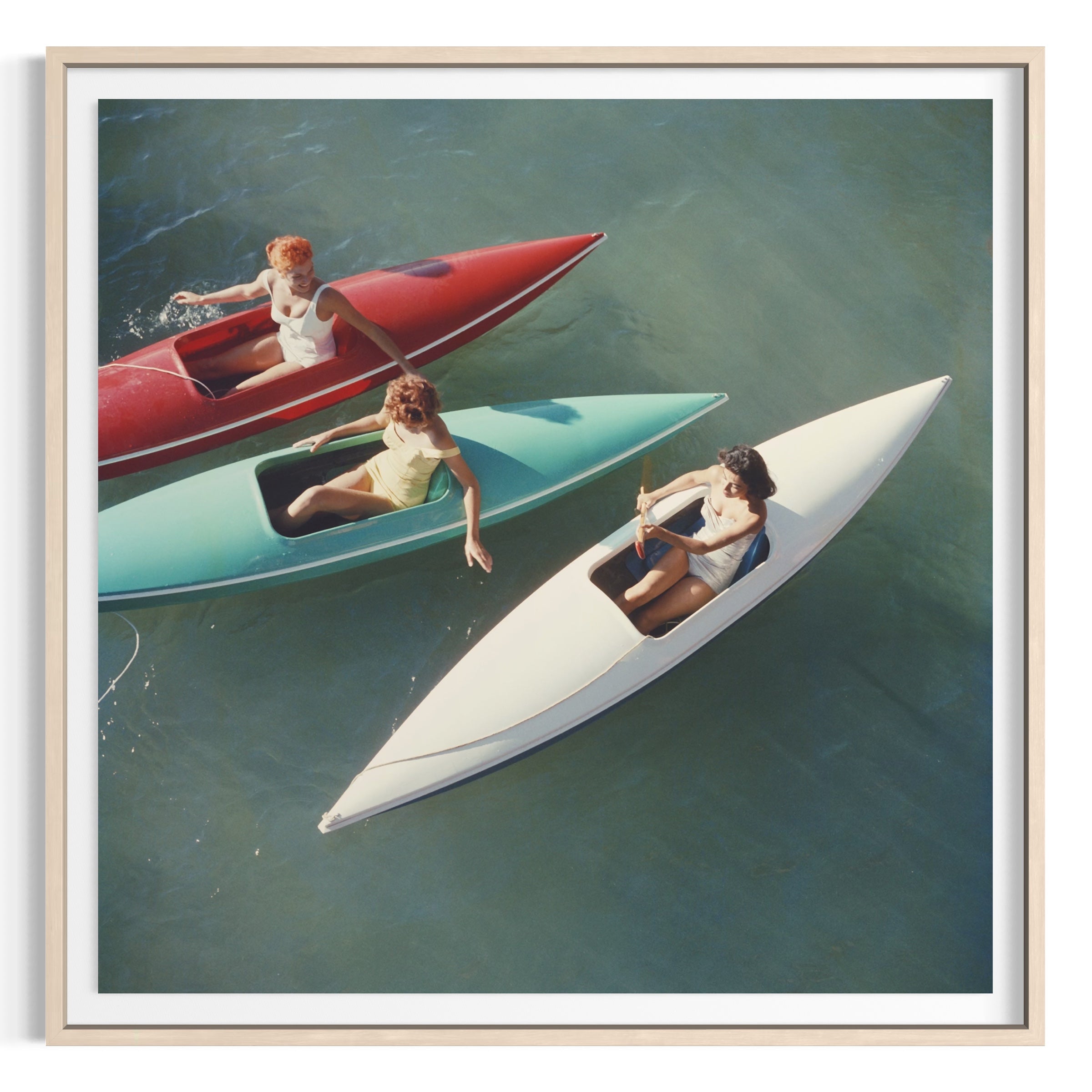 Framed square art print depicting three people in small boats on blue-green water, shown in a light wood frame with a white mat.