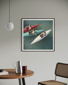 A framed art print depicting three people in small boats on blue-green water, hanging on a light gray wall in a modern room setting with a wooden table, chair, and pendant light visible.