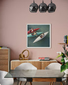 A framed art print depicting two people in colorful rowboats on a teal lake, displayed on a dusty pink wall in a modern dining room setting above a wooden sideboard and dining table.