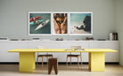 A modern interior scene with a bright yellow table and chairs in front of a white cabinet, above three framed art prints on a light gray wall.