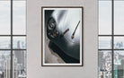 A framed art print depicting a close-up of a silver classic car with a cigar, displayed on a light gray wall between two large windows showing a city skyline.