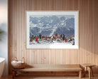 A framed art print depicting a snowy mountain scene with people in winter gear, displayed on a light wood-paneled wall above a wooden bench in a modern interior setting.