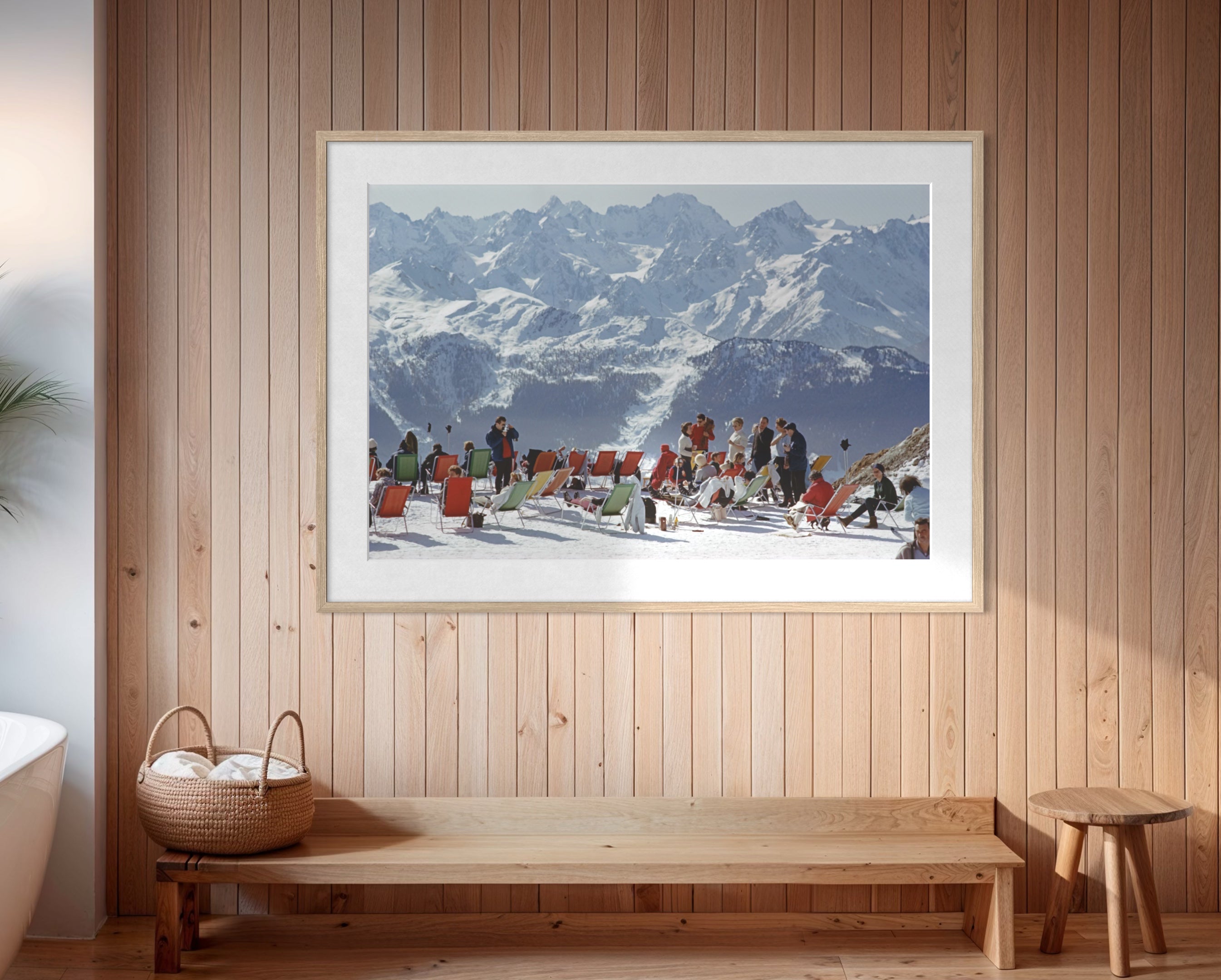 A framed art print depicting a snowy mountain scene with people in winter gear, displayed on a light wood-paneled wall above a wooden bench in a modern interior setting.