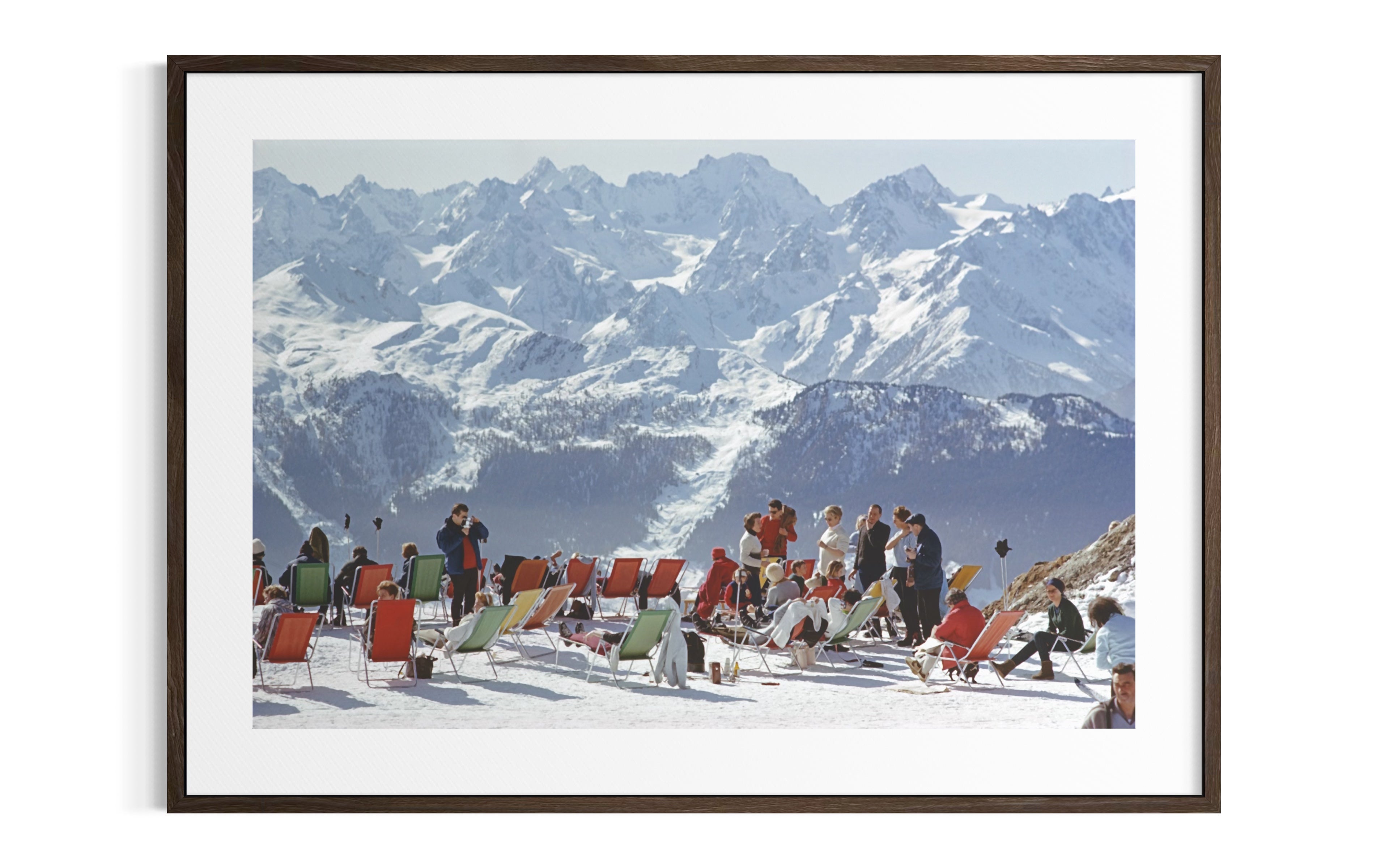 A framed photographic print of a snowy mountain landscape with people sitting in red and green beach chairs in the foreground, shown against a white background.