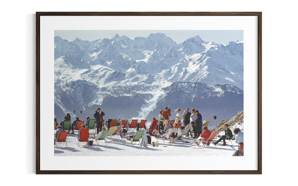 A framed photographic print of a snowy mountain landscape with people sitting in red and green beach chairs in the foreground, shown against a white background.
