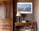 A framed art print depicting a snowy mountain landscape with figures in red clothing, displayed on a brick wall above a wooden desk in a rustic room setting.