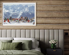 A framed art print depicting a snowy mountain ski resort with people, displayed on a rustic wood-paneled wall above a bed with green pillows and a bedside table.