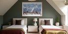 A framed art print depicting a snowy mountain ski resort with colorful skiers, displayed on a dark green wall above two single beds in a modern, symmetrically styled bedroom. The Picturalist Wall Art and Photography for Designed Interiors. Slim Aarons Collection, Skiing. Getty Images Gallery. 