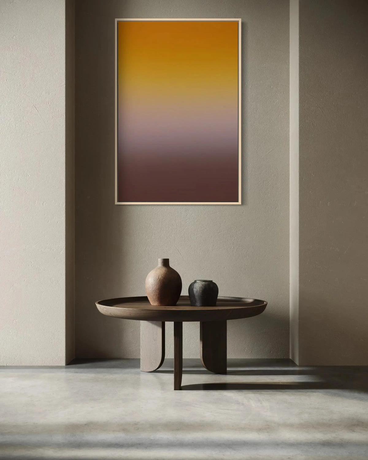 A vertical framed wall art print with a smooth gradient from golden yellow at the top to deep brown and burgundy at the bottom, displayed in a minimalist modern interior above a low round wooden table with ceramic vases.