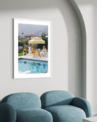 A framed art print depicting a sunny poolside scene with people under a yellow umbrella, displayed on a light gray wall above a blue velvet sofa.