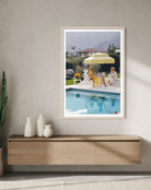 A framed art print depicting a sunny poolside scene with people under a yellow umbrella, displayed on a textured light gray wall above a wooden console table with white ceramic vases.