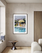 A framed art print depicting a sunny poolside scene with people under a yellow umbrella, displayed on a textured beige wall in a modern room setting.