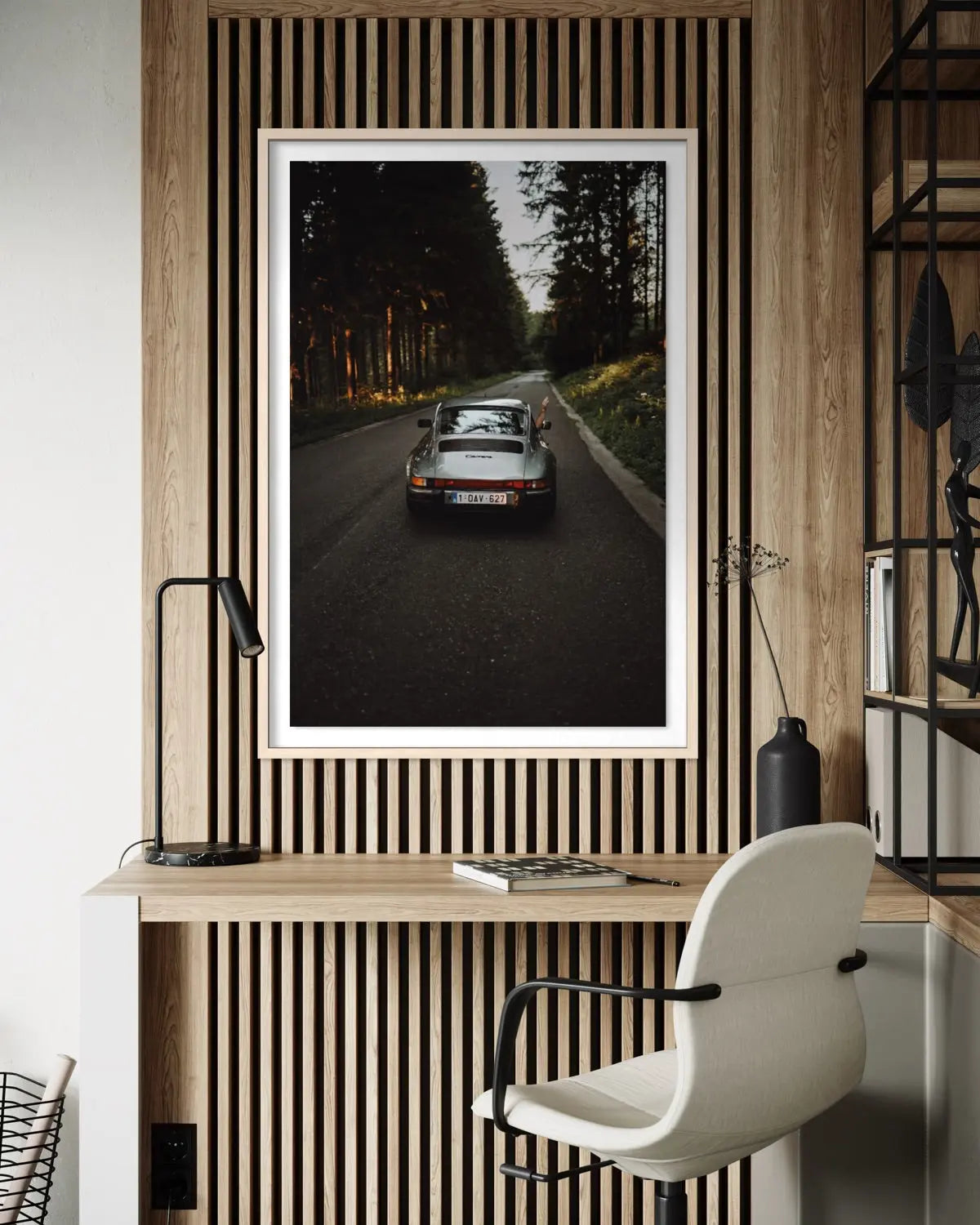 A framed vertical art print depicting a silver sports car on a forest road, displayed on a slatted wood wall above a desk with a chair and lamp.