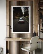 A framed vertical art print depicting a silver sports car on a forest road, displayed on a slatted wood wall above a desk with a chair and lamp.