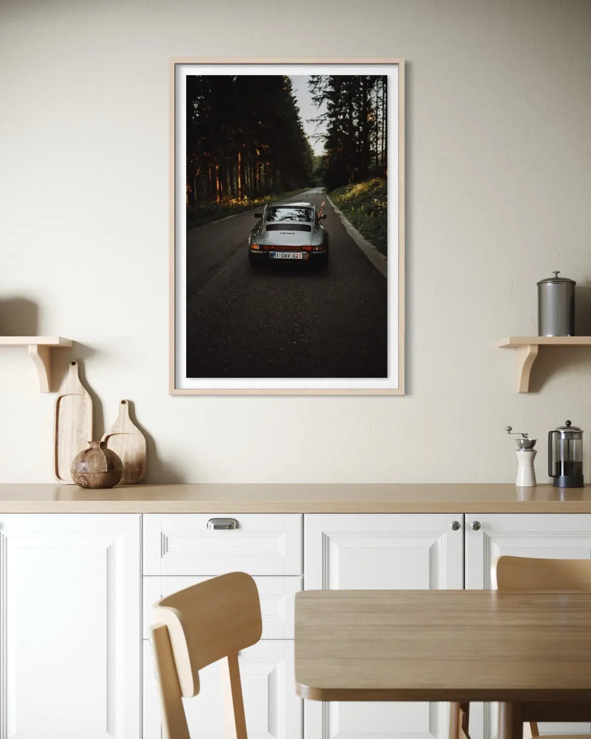 A framed vertical art print depicting a vintage sports car driving down a dark forest road at dusk, displayed on a light beige wall above a modern kitchen counter.