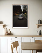 A framed vertical art print depicting a vintage sports car driving down a dark forest road at dusk, displayed on a light beige wall above a modern kitchen counter.
