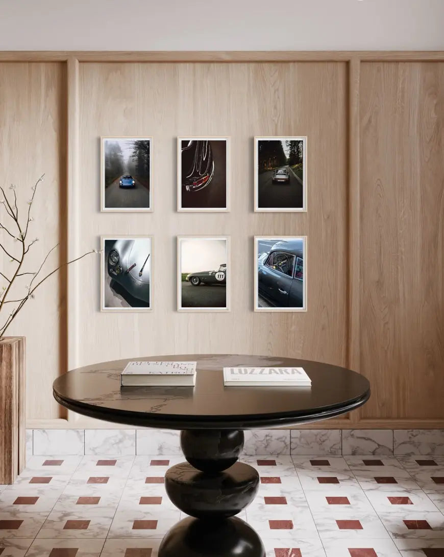 A styled interior scene with a round black table in the foreground and six framed automotive photographs arranged on a light wood paneled wall above. The setting includes a tiled floor and decorative branches at left.