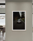 A framed vertical art print depicting a classic sports car driving down a dark forest road, displayed on a neutral beige wall in a modern interior setting.