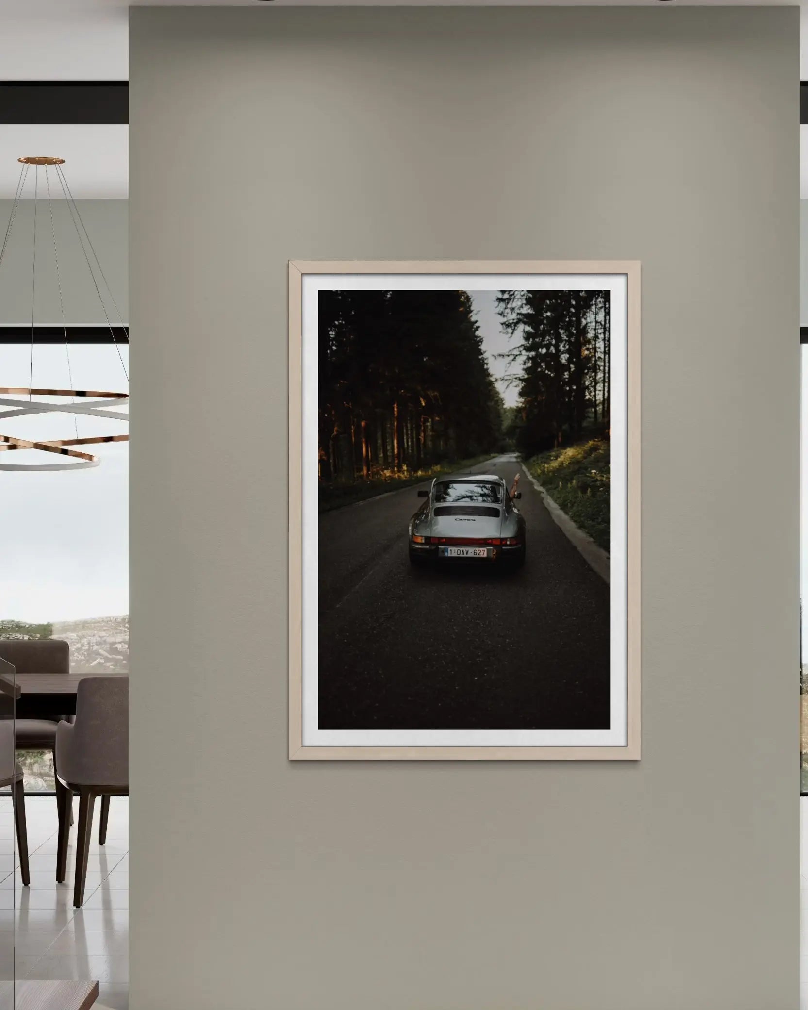 A framed vertical art print depicting a classic sports car driving down a dark forest road, displayed on a neutral beige wall in a modern interior setting.