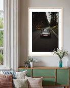 A framed vertical art print of a classic silver sports car driving down a dark forest road, displayed on a beige wall above a green wooden console table with decorative pillows and plants.