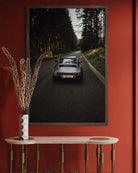 A framed vertical art print depicting a classic Porsche on a forest road, displayed on a red wall above a light wood console table with a vase of dried grass.