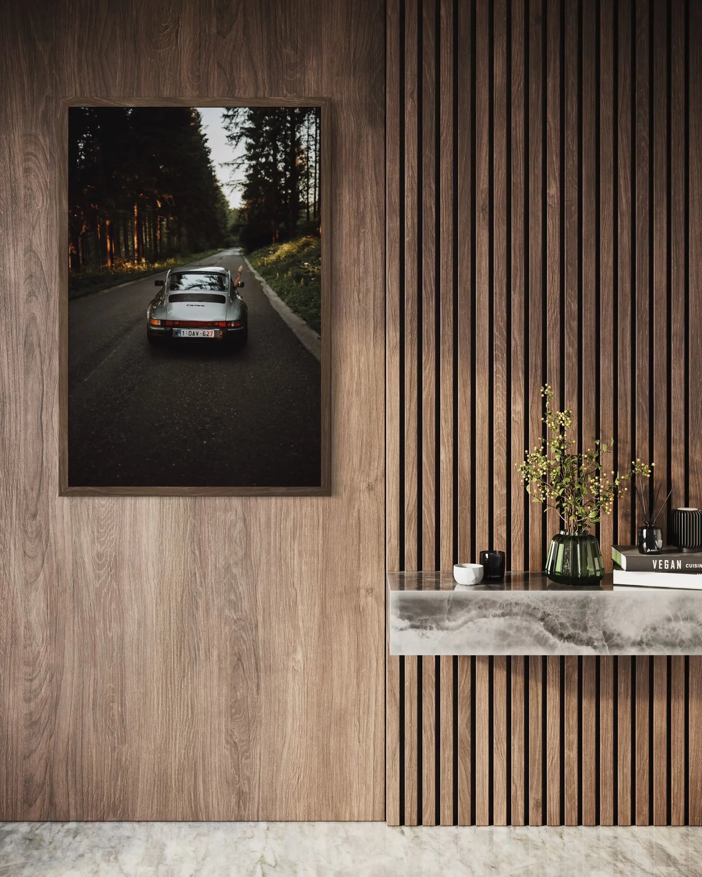 A framed wall art print showing a vintage sports car on a forest road, displayed on a wood-paneled wall above a marble console with decorative objects.