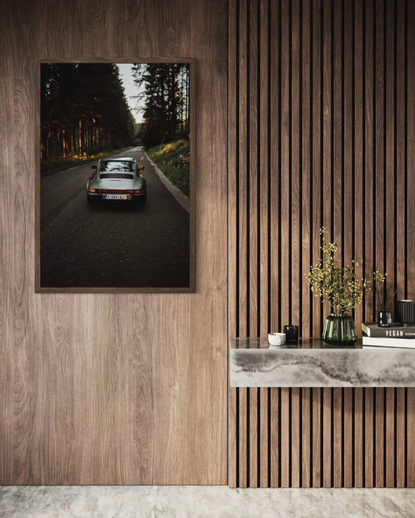 A framed wall art print showing a vintage sports car on a forest road, displayed on a wood-paneled wall above a marble console with decorative objects.