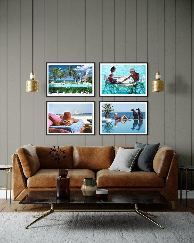 A set of four framed tropical beach and poolside art prints displayed on a gray wood-paneled wall above a brown leather sofa in a modern living room setting.