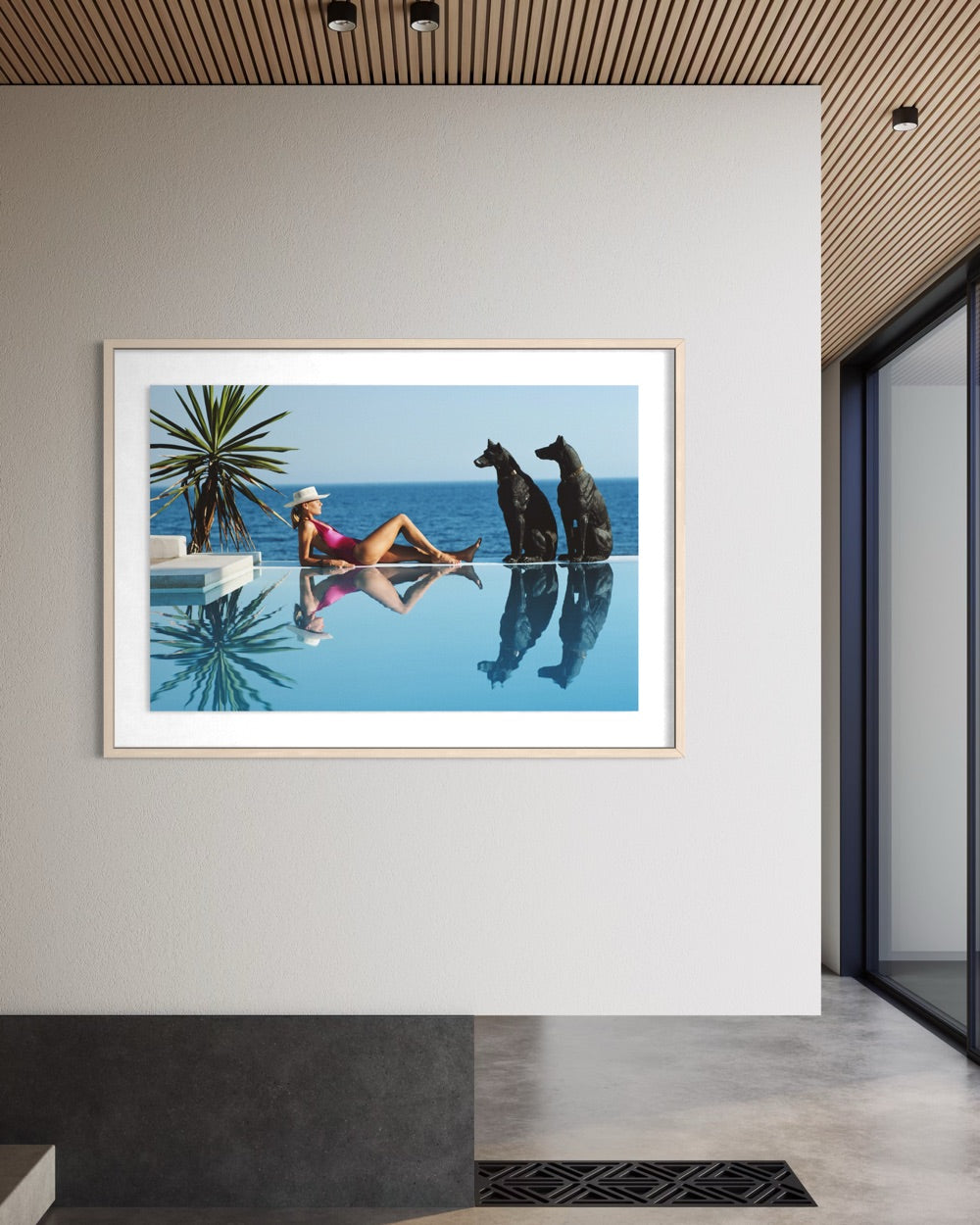 A framed art print depicting a woman in a pink swimsuit lounging by an infinity pool with two black statues, displayed on a light gray wall in a modern interior setting.