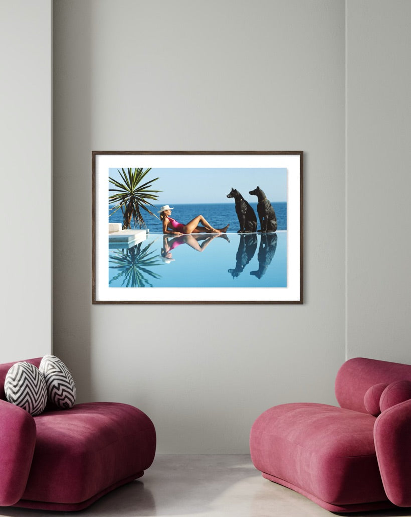 A framed art print depicting a woman in a pink swimsuit relaxing by an infinity pool with two dogs, displayed on a light gray wall above two pink modern armchairs.
