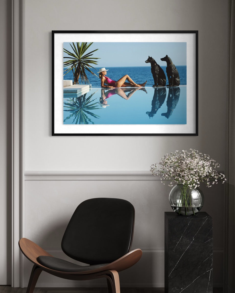 A framed art print featuring a woman in a pink swimsuit sitting on the edge of an infinity pool with two black dogs by the blue ocean, displayed on a gray wall above a modern chair and a marble side table with flowers.