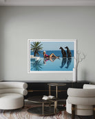 A framed art print depicting a woman relaxing by an infinity pool with two dogs, displayed on a light gray wall in a modern living room setting.