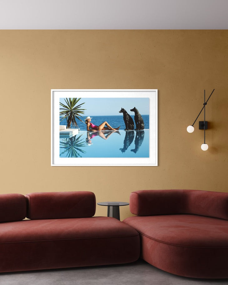 A framed art print depicting a woman relaxing by an infinity pool with two dogs, displayed on a warm beige wall above a modern red sectional sofa.