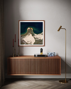 A framed art print depicting a mountain landscape with snow and dark teal sky, styled on a gray wall above a wooden console table in a modern room setting.
