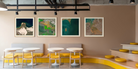 Four framed landscape art prints with white frames hang on a beige wall in a modern interior setting with curved yellow seating, white tables, and industrial ceiling details.