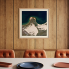 A framed art print featuring a dramatic aerial view of a snow-capped mountain peak meeting deep blue water, displayed on a wood-paneled wall above a dining table with three tufted rust-orange chairs.