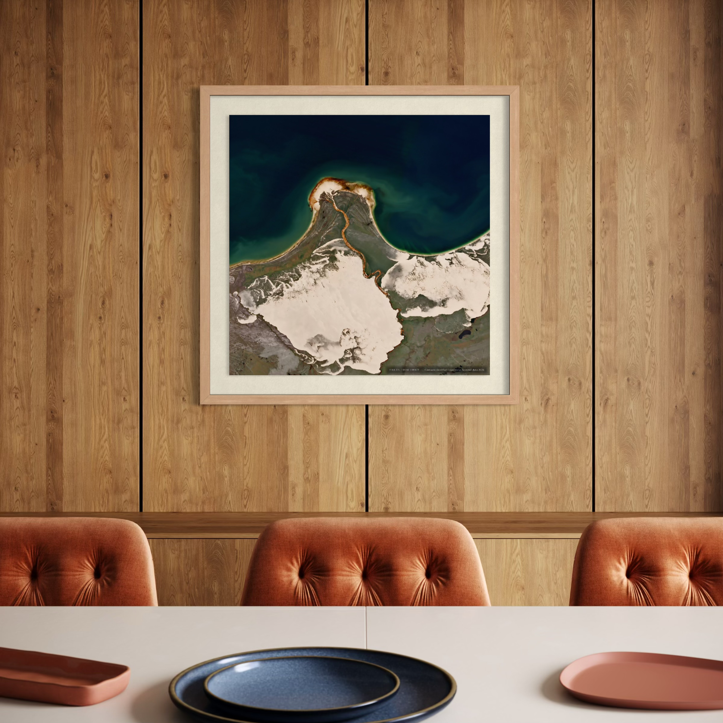A framed art print featuring a dramatic aerial view of a snow-capped mountain peak meeting deep blue water, displayed on a wood-paneled wall above a dining table with three tufted rust-orange chairs.