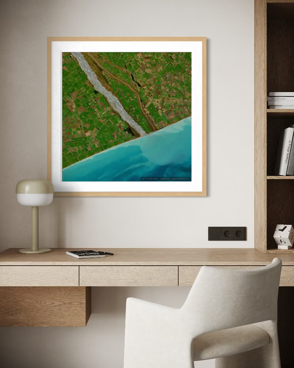 A framed aerial landscape print of a green field with a winding river and turquoise water, displayed on a light beige wall above a wooden desk with a chair and lamp.