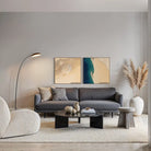 A modern living room featuring a two-panel abstract wall art set hanging above a dark gray fabric sofa. The artwork has soft beige, cream, and teal-blue abstract shapes.