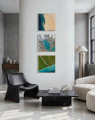 Three framed abstract landscape art prints with teal, green, beige, and gray tones are displayed in a vertical row on a white wall in a modern living room setting.