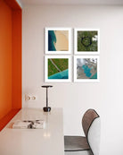 Four framed abstract landscape prints arranged in a 2x2 grid on a white wall above a desk in a modern room with an orange accent wall and a gray chair.