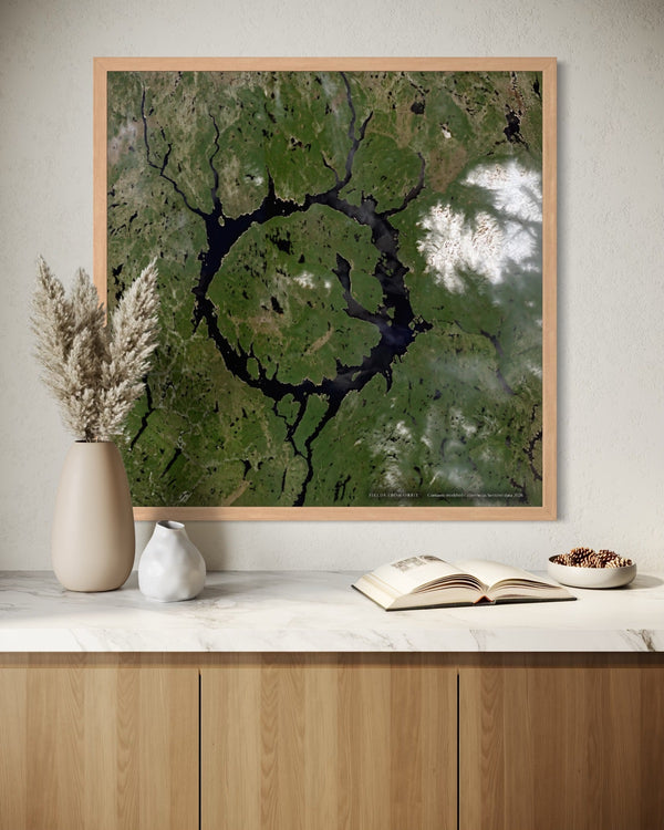 A framed square art print featuring a green aerial landscape with a dark ring-shaped lake, displayed on a light wall above a marble countertop with decorative vases, an open book, and a small bowl.