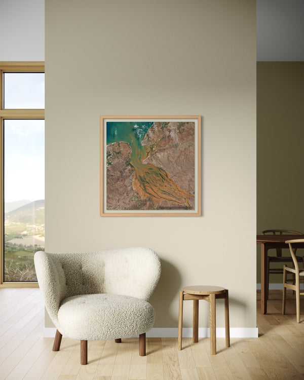 A framed art print featuring an aerial view of a winding river through a valley, styled in a modern living room setting above a cream-colored armchair and a small wooden stool.