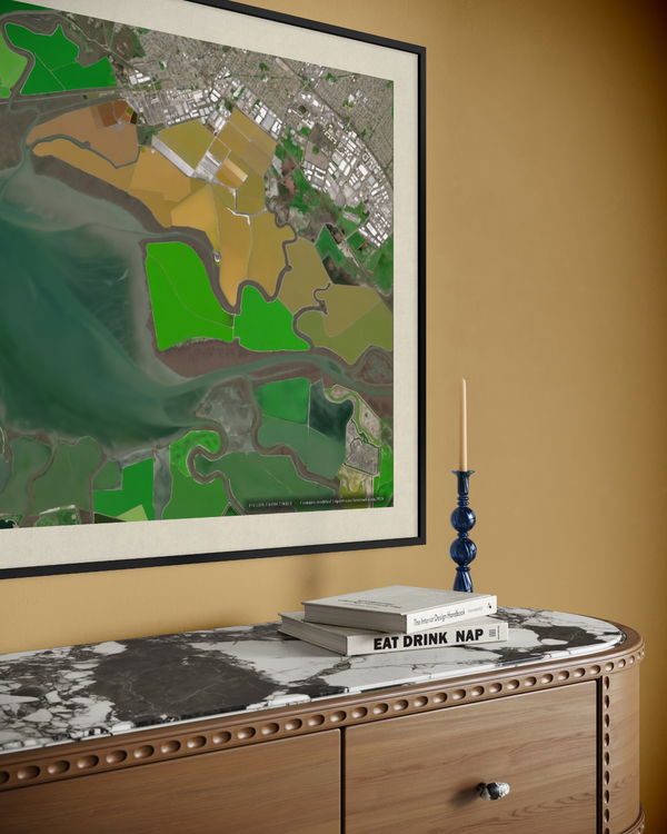 A framed aerial map print featuring green agricultural fields and waterways, displayed on a warm mustard wall above a wooden console table with a marble top and decorative books.